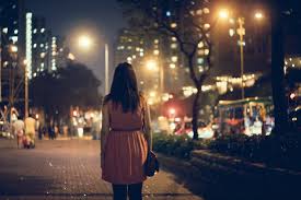 A woman walking at night. You can see her back and city lights around her.